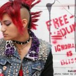 Eine Punkerin mit rotem Iro und Jeansjacke vor einer weißen, besprayten Wand (Freedum/Ignorance is bliss)