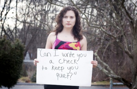 Eine Frau hält ein Schild hoch mit der Aufschrift: Can I write you a check to keep you quiet?