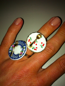 Foto einer Hand mit zwei Fingerringen, die mit kleinen Tellern und Besteck dekoriert sind.