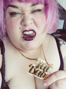 Porträtaufnahme einer Person mit Make-Up und bunt gefärbtem Haar, die eine goldene Halskätte trägt. Der Anhänger bildet den Schriftzug"Fat babe".